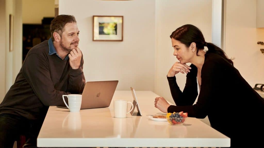 A man and woman talk at a kitchen island, with a laptop and fruit.