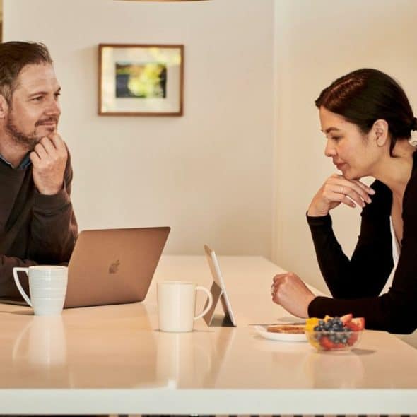 A man and woman talk at a kitchen island, with a laptop and fruit.