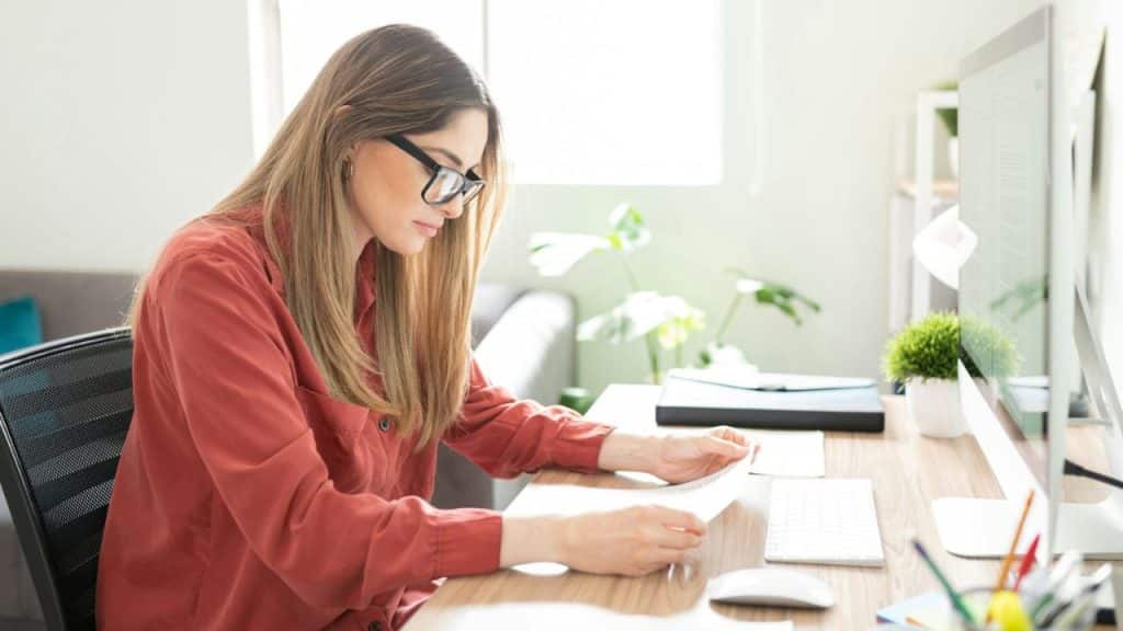 A woman with long brown hair and glasses sits at a desk, looking down at a document in her hands.