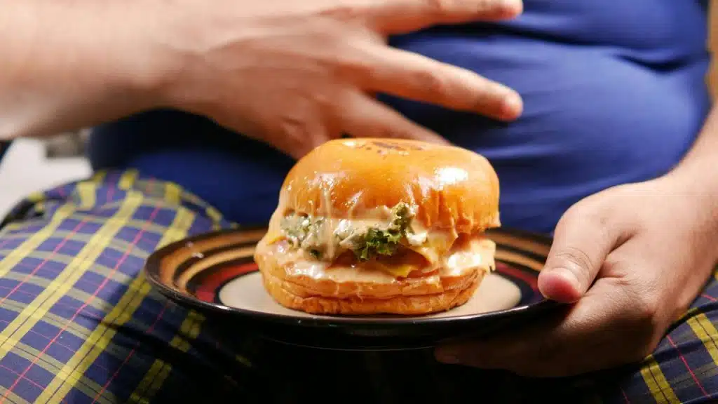 A saucy cheeseburger with melted cheese and lettuce on a plate held before a person’s prominent belly.