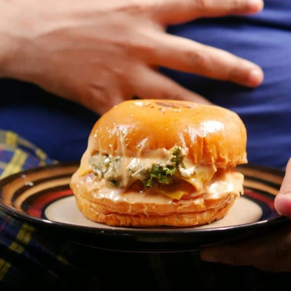 A saucy cheeseburger with melted cheese and lettuce on a plate held before a person’s prominent belly.