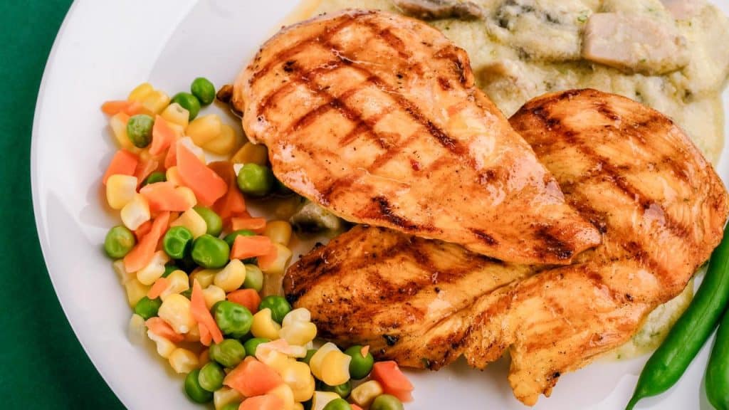 A plate of grilled chicken breasts with peas, carrots, corn, and creamy mashed potatoes.