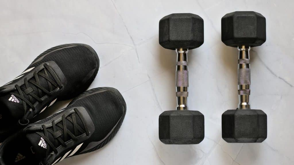 A pair of black Adidas sneakers beside two black hex dumbbells on a white marble surface.