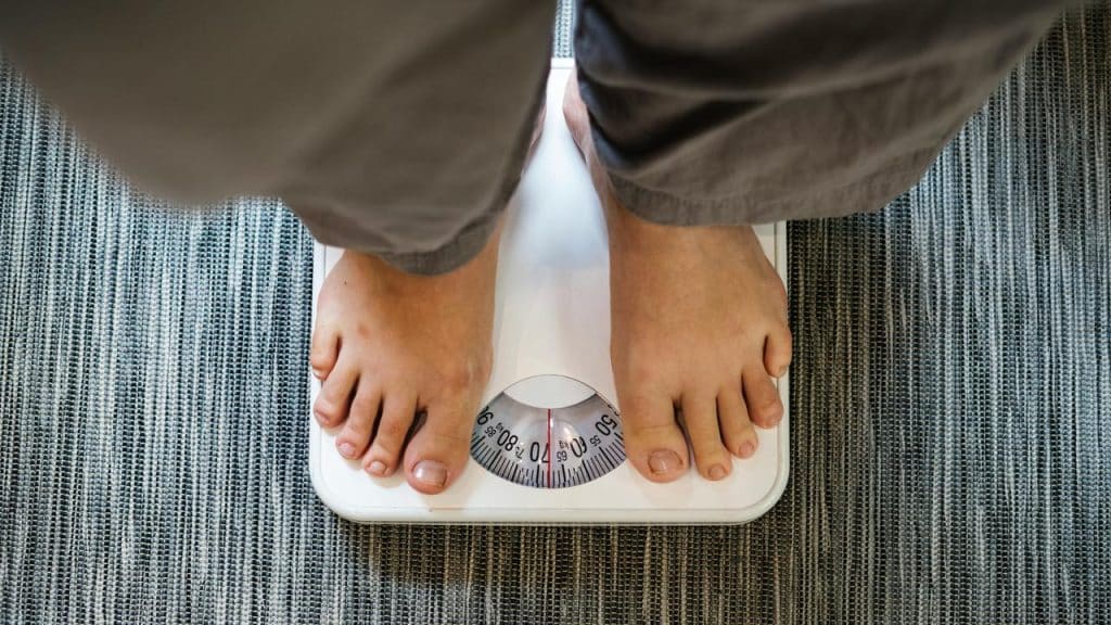 A person’s bare feet standing on a white analog bathroom scale atop a gray woven rug.