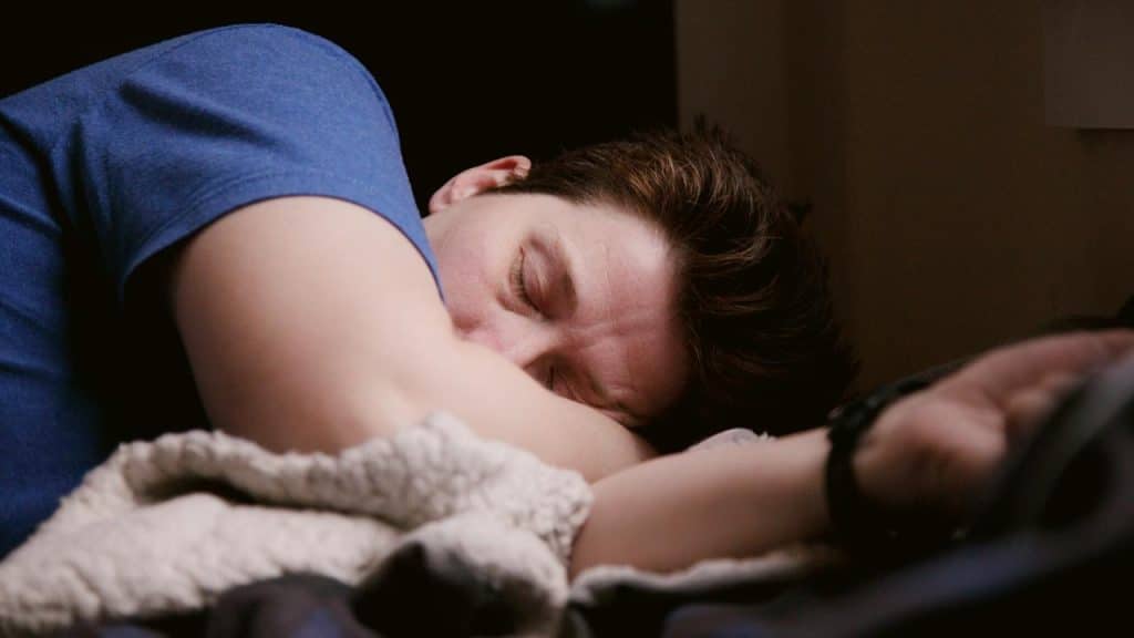 A person in a blue shirt asleep with their head resting on folded arms over a soft blanket.