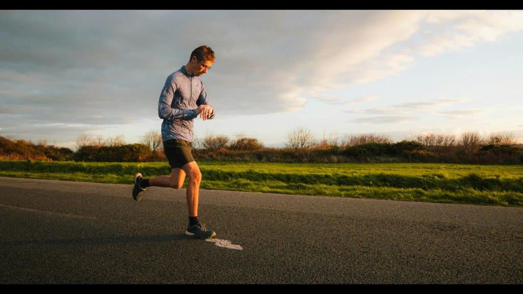 A runner in a blue top glancing at a smartwatch while jogging on a road beside a grassy field.