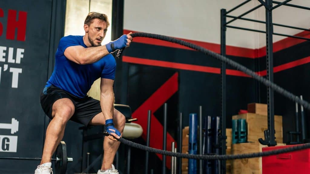 A man in a blue shirt powerfully swinging heavy battle ropes in a gym.