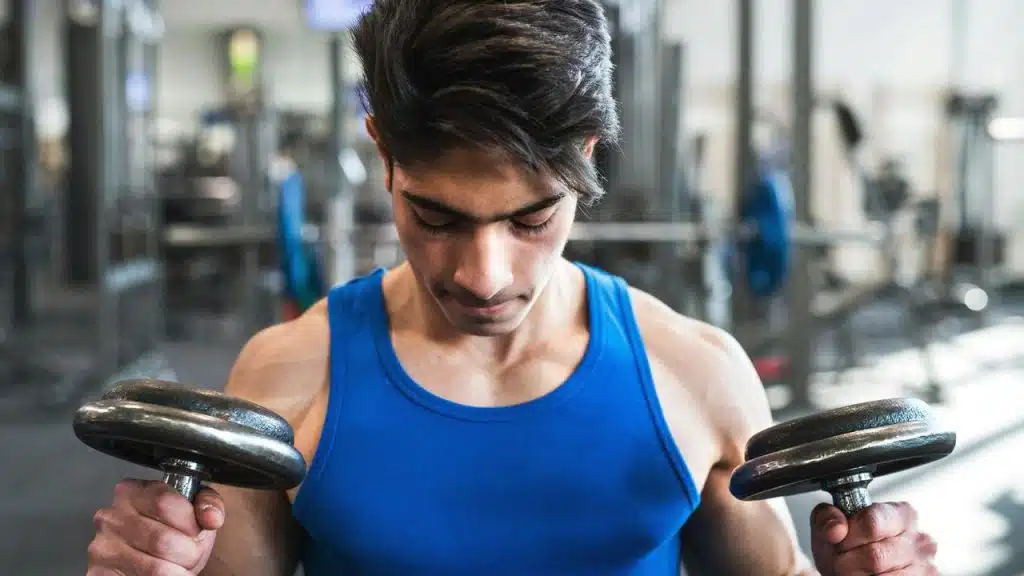 A young man in a blue tank top lifting dumbbells in a gym.