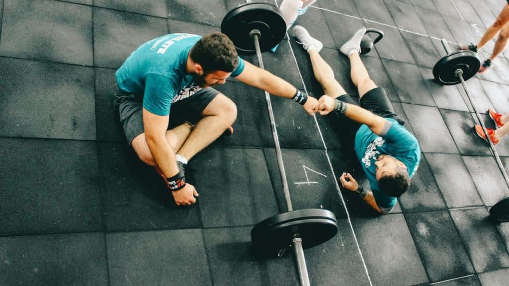 A kneeling man fist-bumps a man lying next to a barbell on a gym floor.