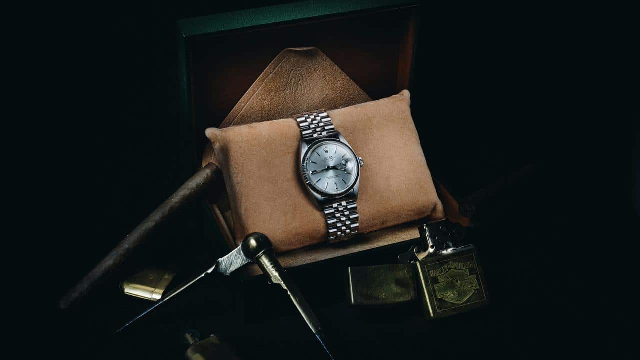 A silver wristwatch resting on a suede cushion inside a display box, surrounded by a cigar, lighter, and brass tools.
