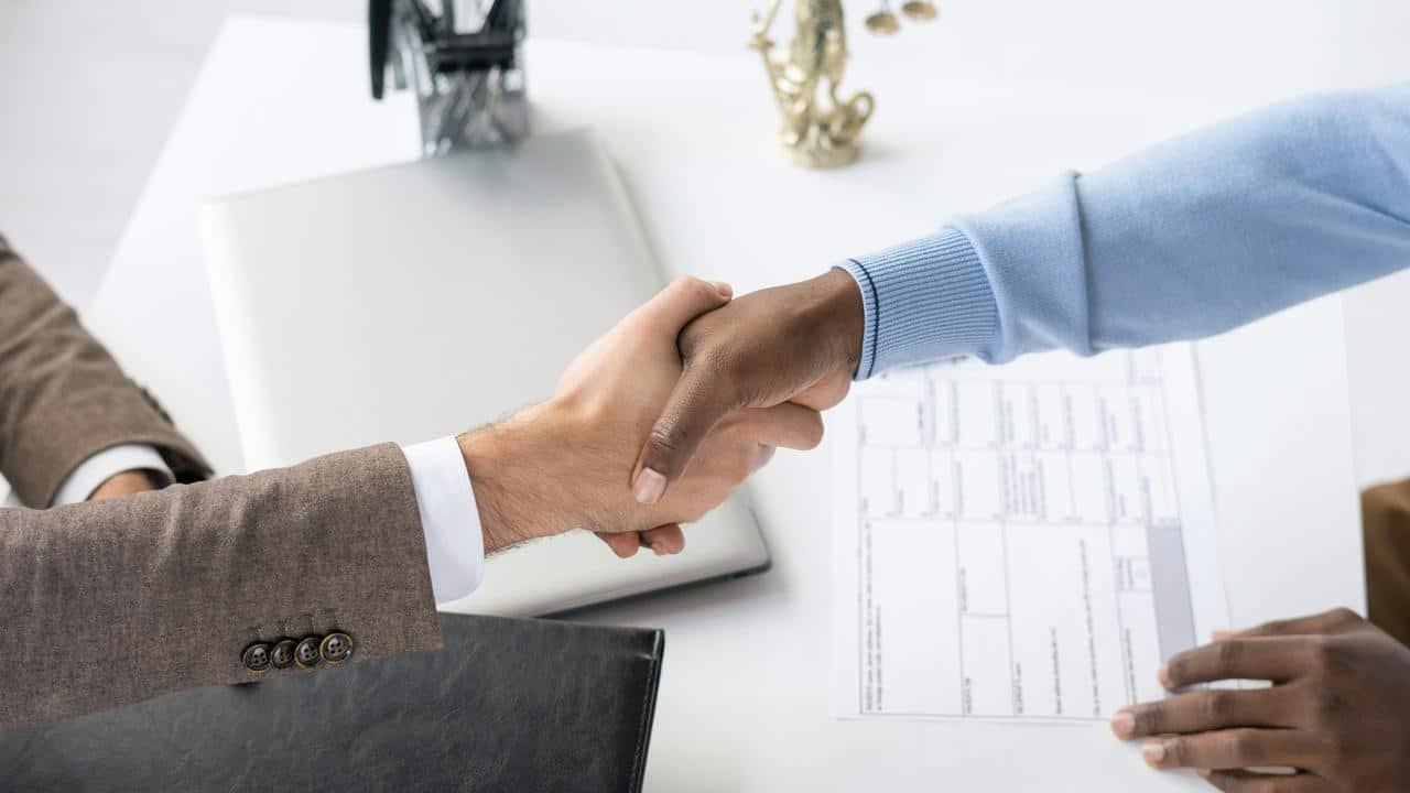A handshake over a desk with documents and a laptop nearby.
