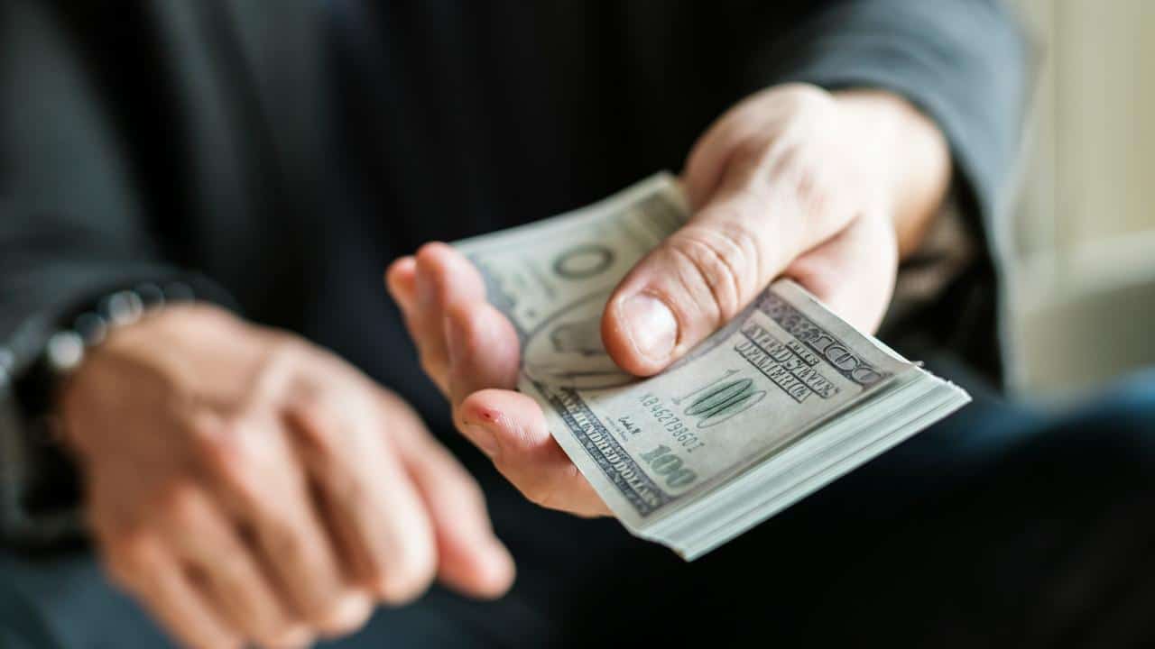 A close-up of a hand offering a stack of hundred-dollar bills.