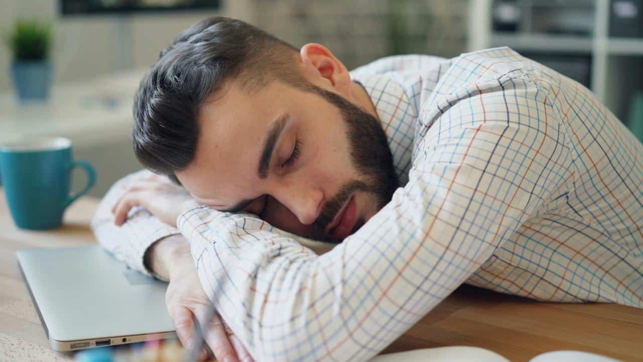 A man in a checked shirt sleeping at his desk with his head resting on his arms.