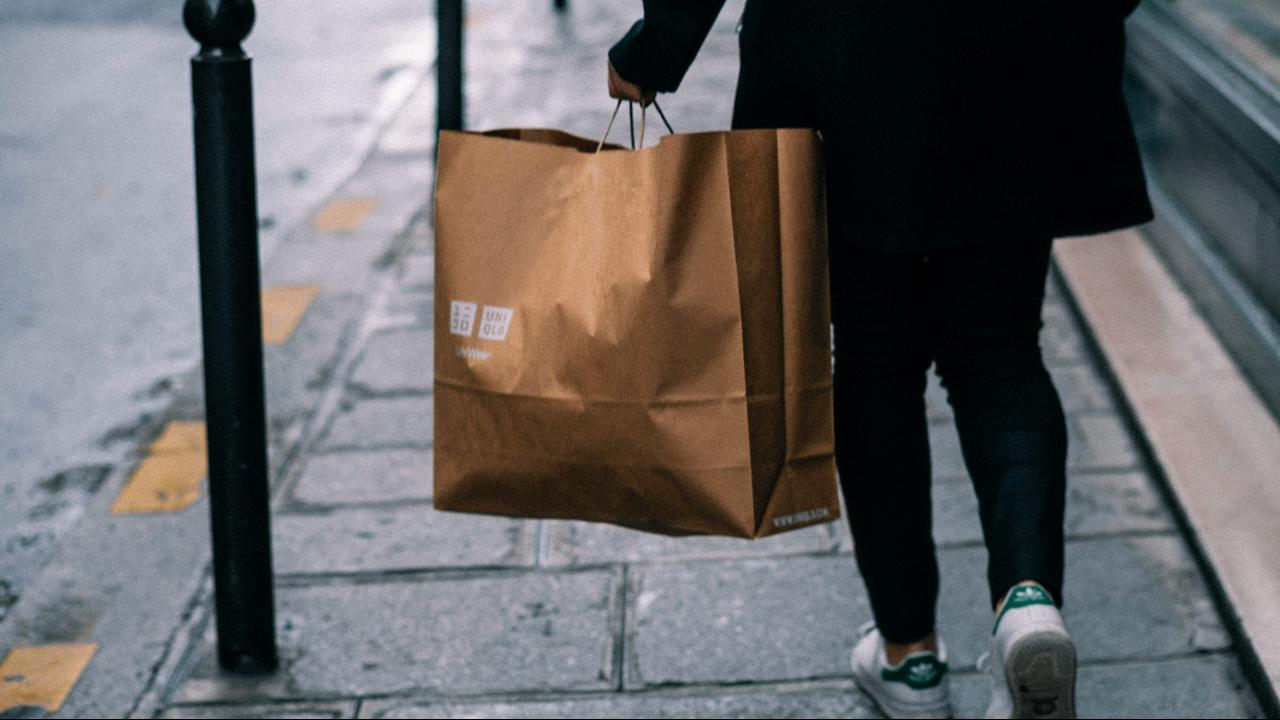 A person walking on a sidewalk holding a brown Uniqlo shopping bag.