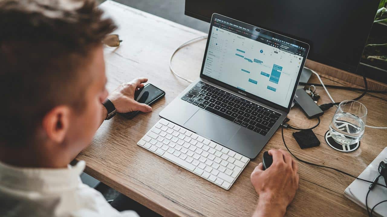 A person at a desk viewing a calendar on a laptop while holding a phone and using a mouse.