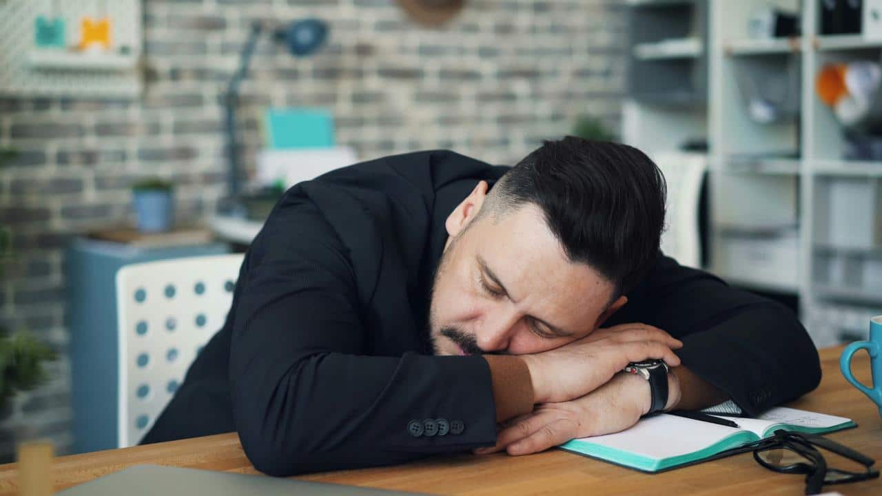 A man in a suit sleeping at his desk with his head resting on folded arms over an open notebook.