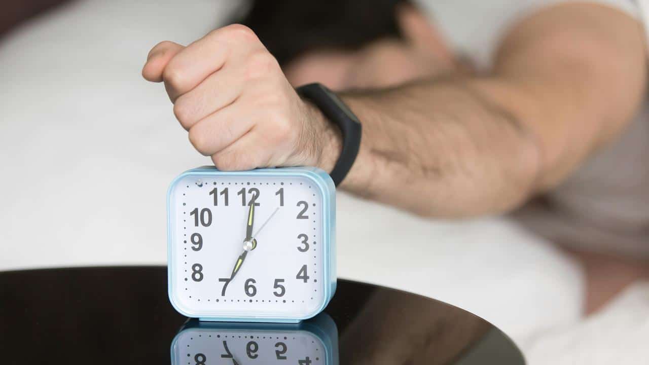 A fist pressing down on a light blue alarm clock on a bedside table.