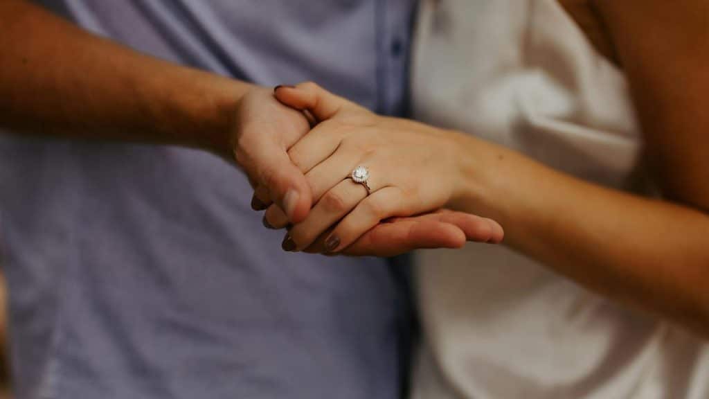 A woman’s hand wearing a diamond engagement ring held in a man’s hand.
