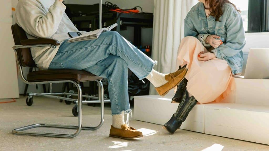 A man in jeans and loafers and a woman in a skirt and cowboy boots seated and talking in a studio space.