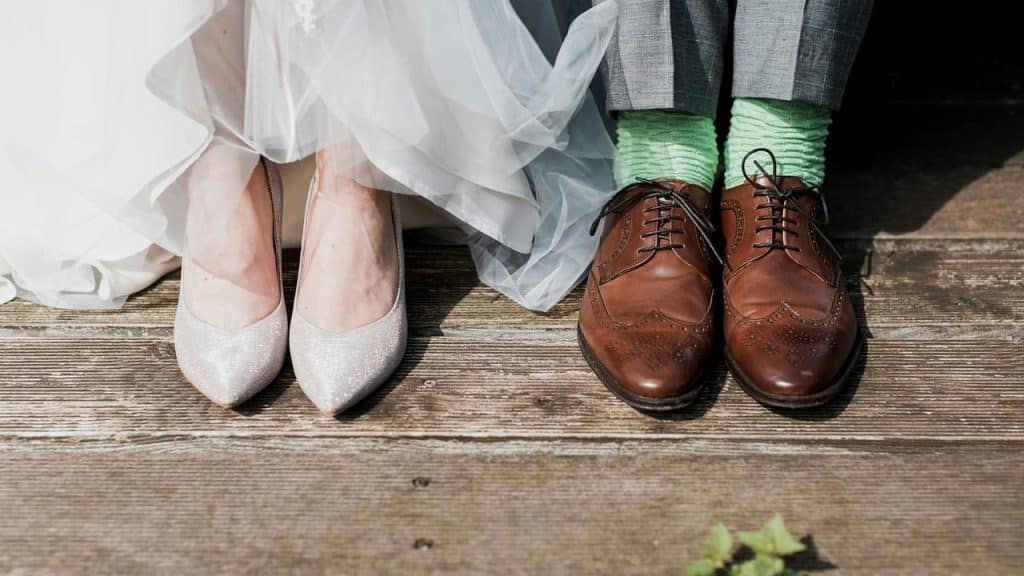 A pair of glittery women’s heels next to brown brogues with bright green socks on wooden planks.