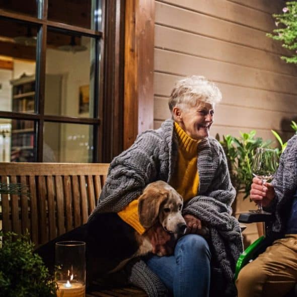 An elderly couple and a dog sit outdoors at night, with the man holding a wine glass.