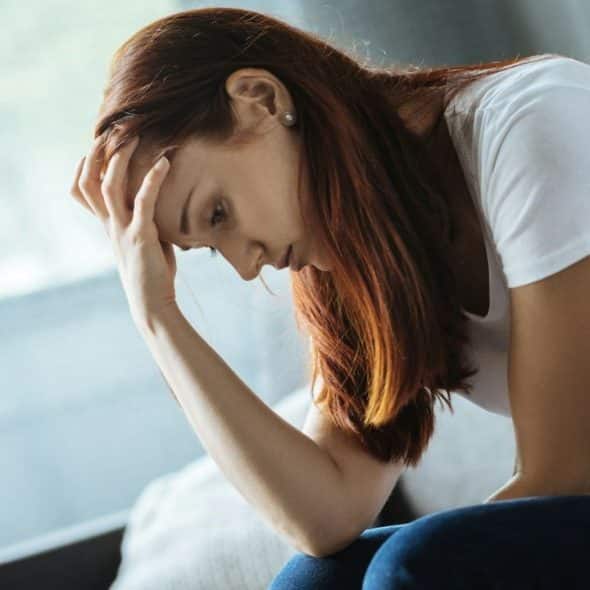 A red-haired woman sits, looking down with one hand on her forehead.