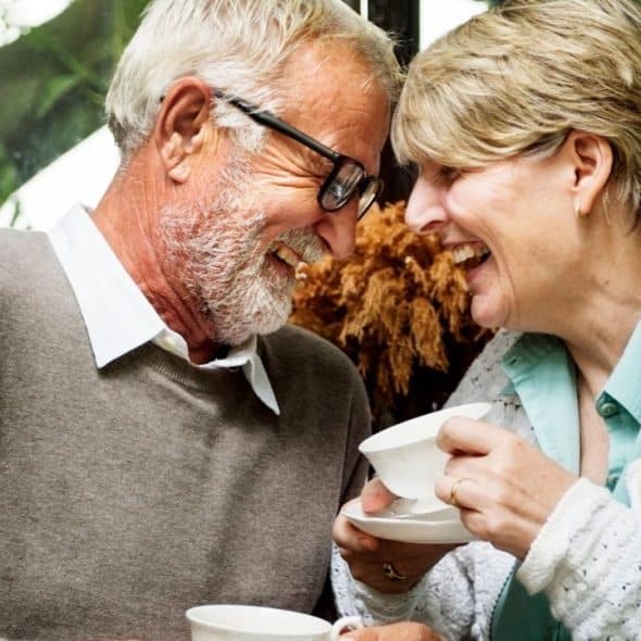 A happy older couple with grey hair smiles and laughs together over coffee cups.