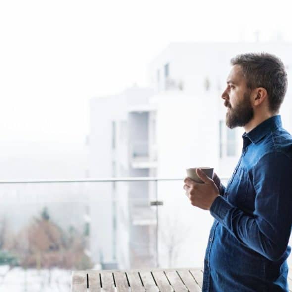 A bearded man holds a cup on a balcony, looking out at buildings.