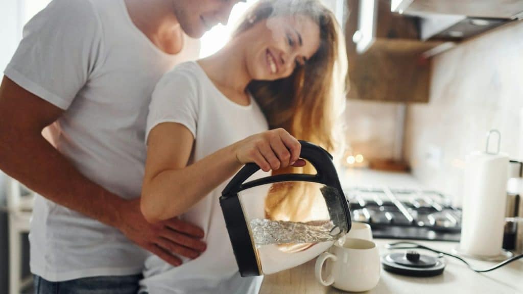 A man hugs a smiling woman pouring water from a kettle into a mug.