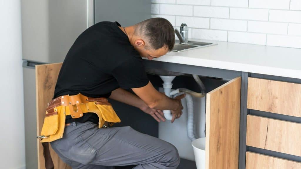 A man wearing a tool belt is crouching and working on a pipe under a kitchen sink.