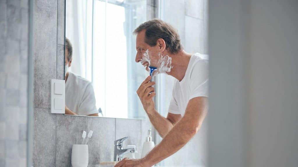 A man with a lathered face shaves with a blue razor in a bathroom mirror.