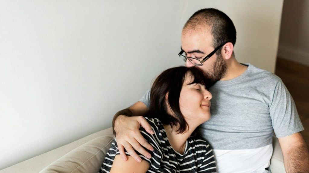 A man wearing glasses gently kisses a woman's forehead while embracing her.