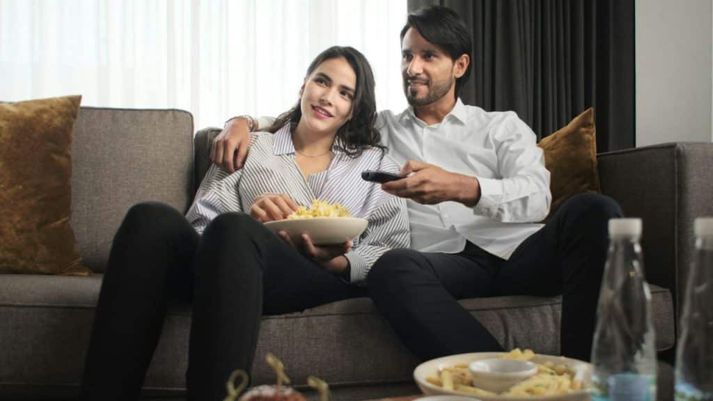 A smiling couple sits on a couch, watching TV and eating popcorn.
