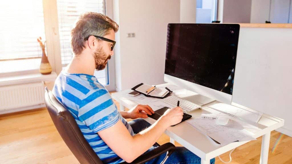 A man with glasses and a beard works at a desk using a drawing tablet.