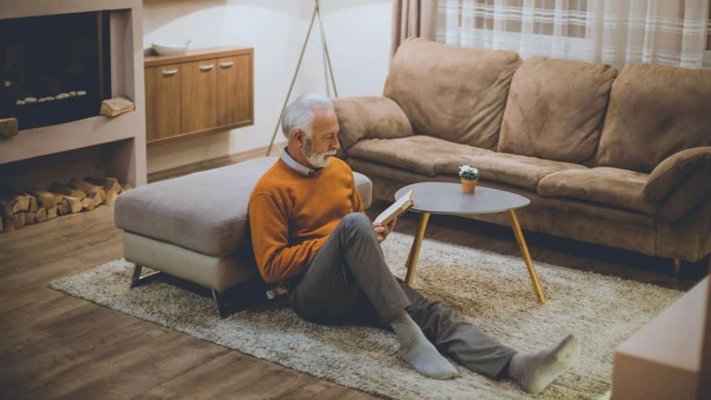 A senior man in an orange sweater sits on the floor, reading a book.
