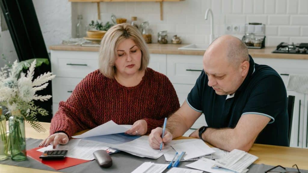 A mature couple reviews documents and calculates finances at a kitchen table.