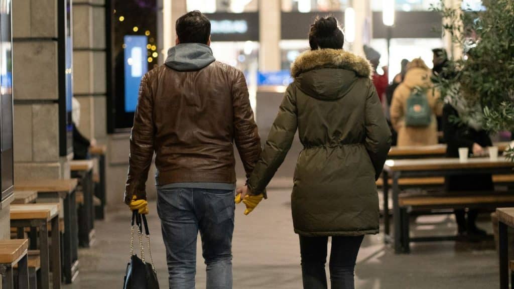 A couple holding hands walks away from the camera through an outdoor market.
