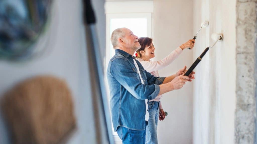 A happy older couple smiles while painting a wall with rollers.
