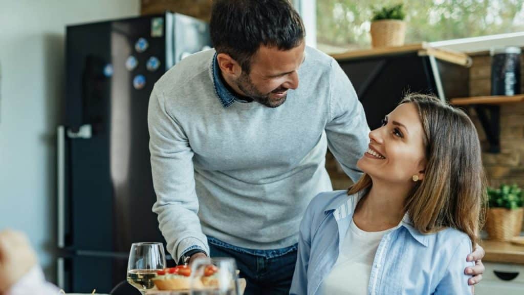 A smiling man stands behind a woman, looking at her with affection.