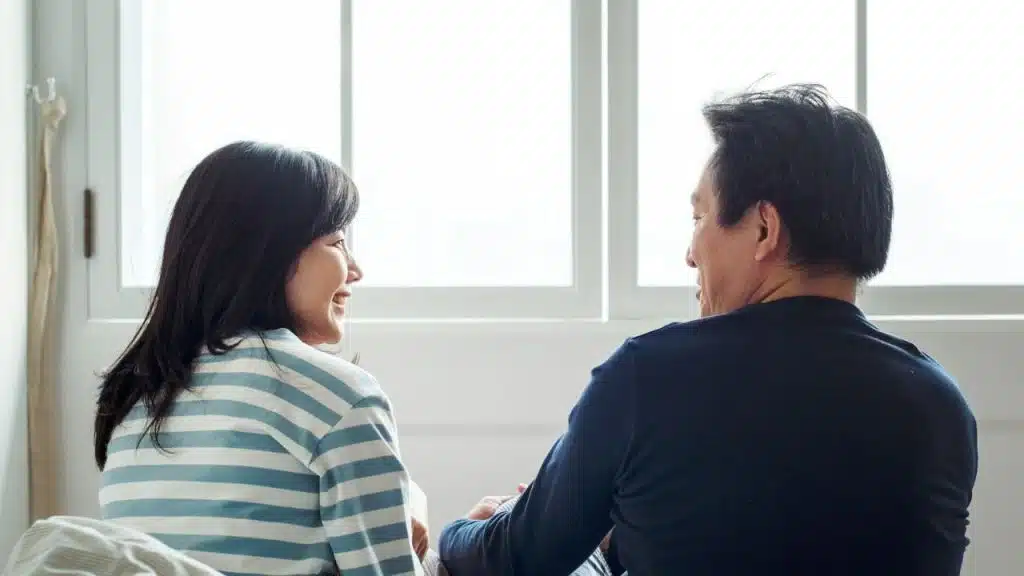 A man and a woman are seated facing each other, looking happy, with a bright window behind them.