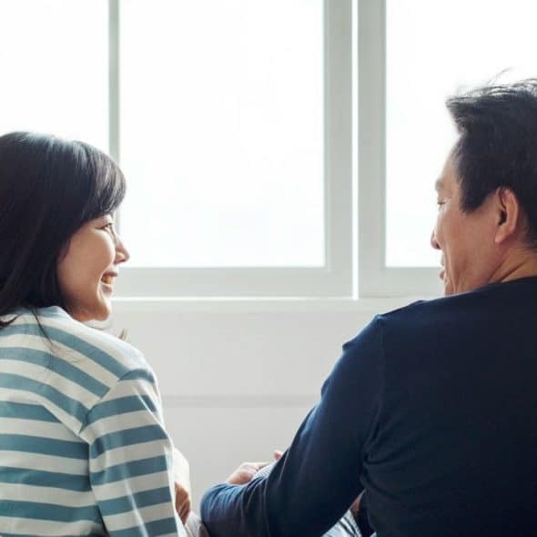 A man and a woman are seated facing each other, looking happy, with a bright window behind them.