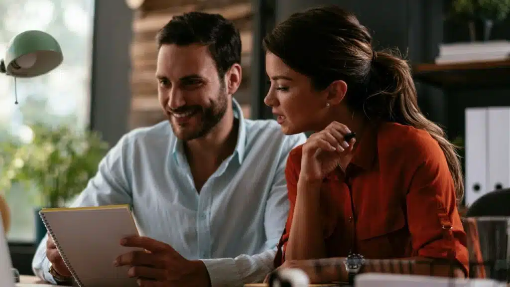 A smiling man and woman look at a notebook together at a desk.