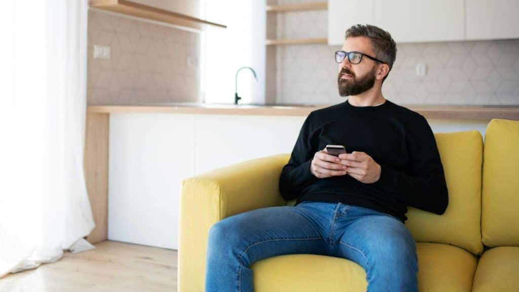 A thoughtful man with a beard and glasses sits on a yellow couch holding a phone.