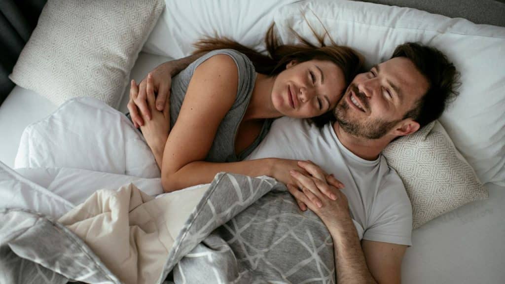 A happy couple lies in bed, embracing and smiling with eyes closed.