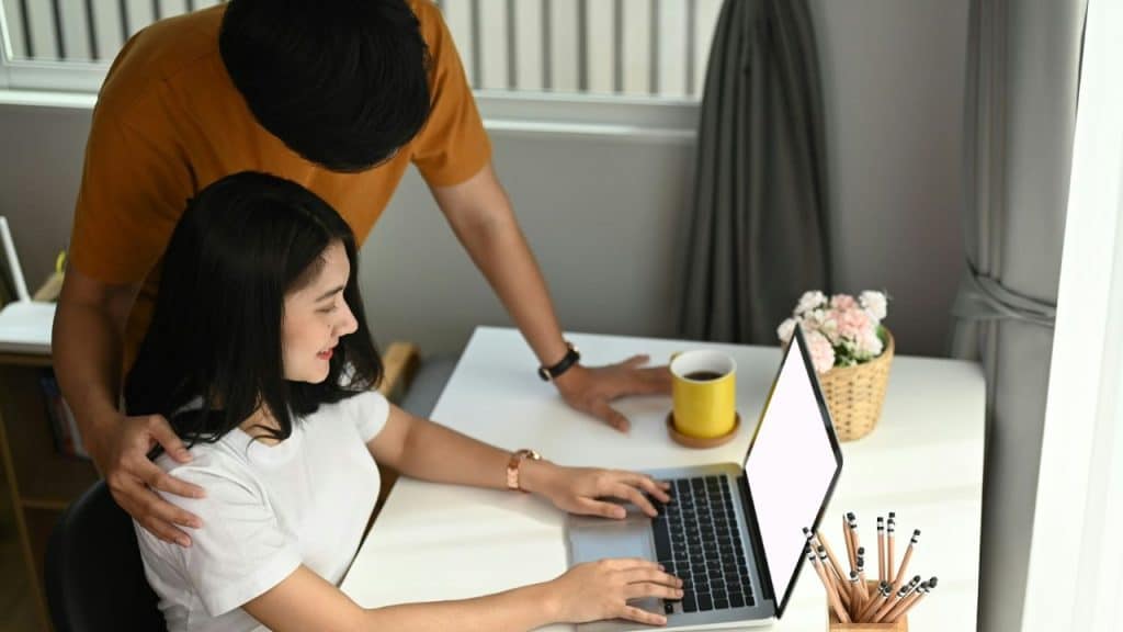A woman works on a laptop at a desk while a man stands behind her.