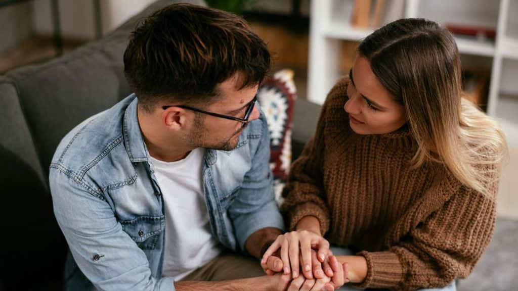 A man and a woman sit close, holding hands and looking at each other intently.