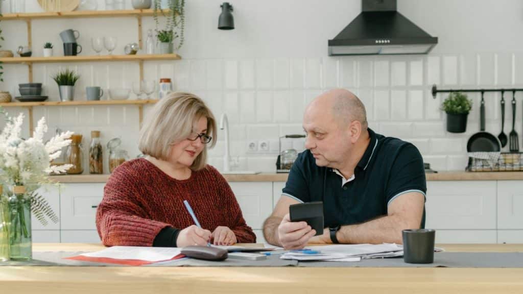 A man holds a calculator and talks to a woman writing at a kitchen table.