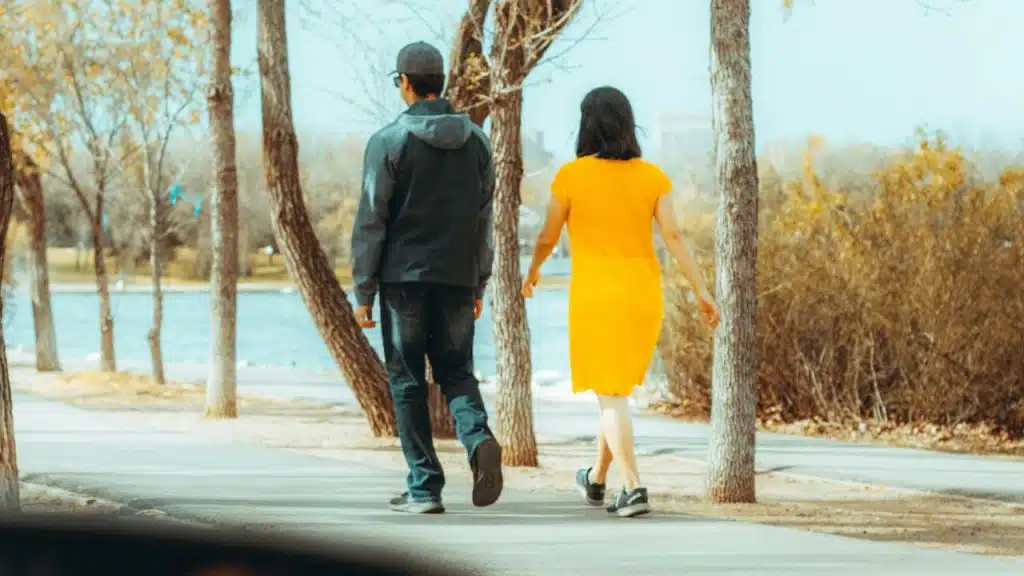 A man and woman walk along a path beside a lake on a sunny day.
