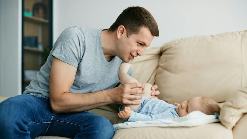 A smiling father changes his baby's diaper on a couch.