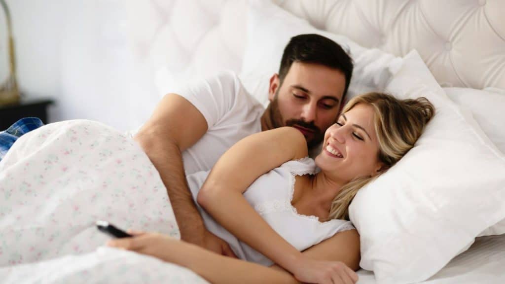 A couple is lying in bed, with the woman smiling and holding a remote control.